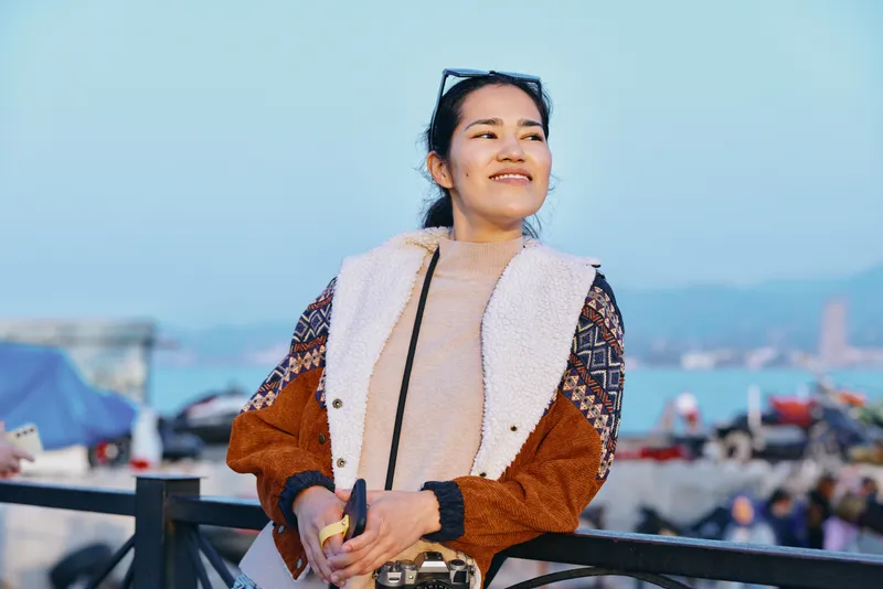 Outdoors, smiling woman in a cozy jacket poses on a boardwalk with sunglasses, capturing a casual fashion moment and relaxed lifestyle under clear sky and bright light, conveying confidence and