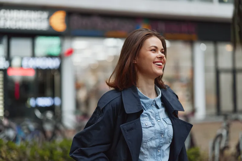 Young woman with brown hair smiles happily while walking outdoors in casual clothes during the day in an urban environment.