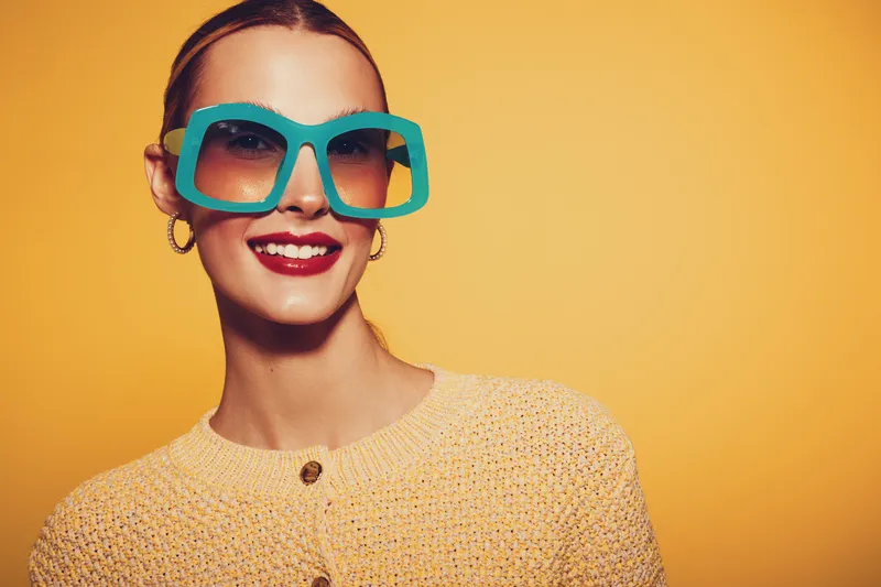 Smiling woman with large turquoise sunglasses and red lipstick wears yellow knitted sweater against bright yellow background. Concept of fashion, style, summer, and cheerful mood.