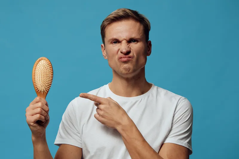 Man holding hairbrush with a funny, confused facial expression, pointing at it while wearing a plain white t-shirt on vibrant blue background. Lifestyle, humor, casual concept, studio shot, emotional,