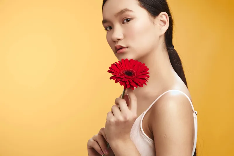 asian woman holds a red flower close to her face in a studio portrait, warm orange background, soft lighting, conveying calm elegance and a contemporary beauty vibe.
