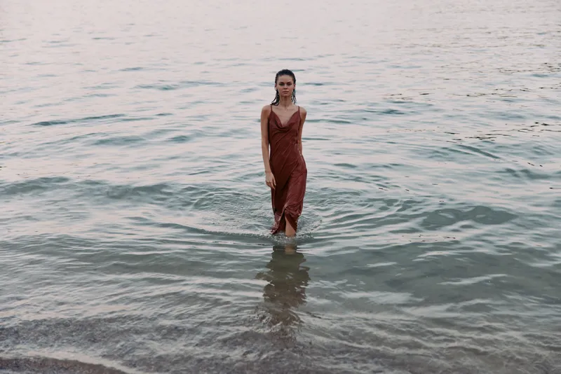 Woman wearing a red dress stands in shallow sea water, creating a calm, cinematic focal point as gentle waves lap at her feet and a soft horizon glows.