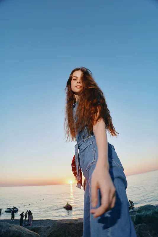 lifestyle woman in modern boho-western denim street style with long hair by the ocean at sunset, film color capturing relaxed summer vibes and natural beauty