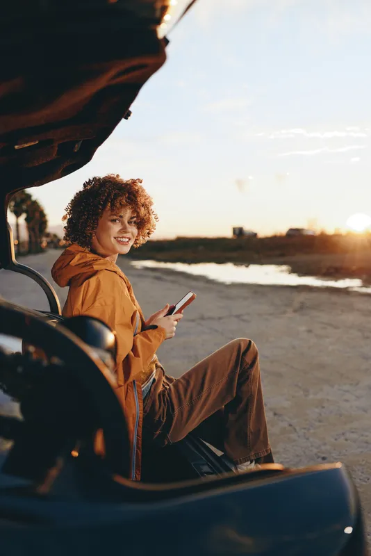 Woman lifestyle on beach at sunset, sitting near car door, smiling and holding phone, casual travel outfit, curly hair, warm light, sunset glow, road trip outdoors, coastal evening