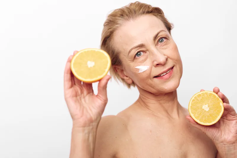 Mature woman with natural skin care cream under eyes holding two halves of fresh orange. Concept of healthy aging, organic beauty, wellness, and vitamin c skincare routine in bright studio.