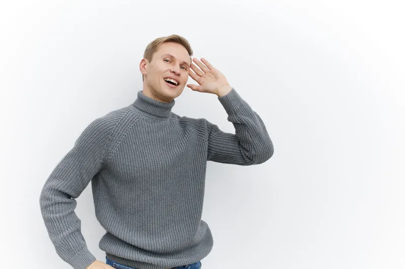 young man smiling cheerful gray knitted sweater standing bright white background hand near ear digital detox concept