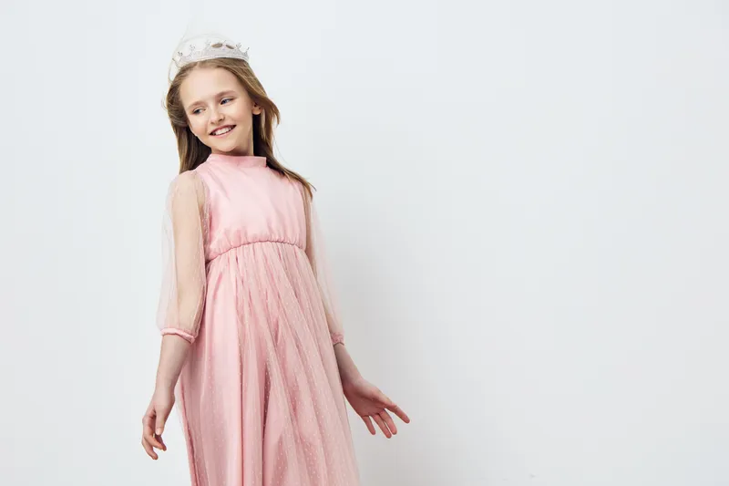 A joyful young girl in a light pink dress with sheer sleeves, smiling gracefully against a soft white background, embodying innocence and playfulness