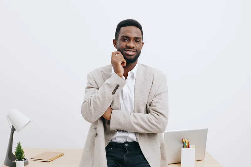 Professional confident smiling young African American man in casual blazer stands in office studio with modern workspace background, holding chin with hand, looking at camera. Business success concept