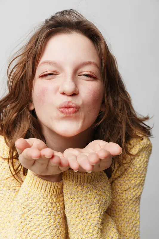 Young woman with wavy hair wearing yellow sweater blowing a kiss with closed eyes. Studio portrait on white background. Concept of affection, friendliness, and positive emotions.