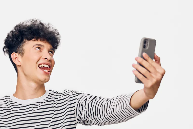smiling man taking selfie with smartphone wearing striped shirt against white background, showing joyful expression and modern casual style