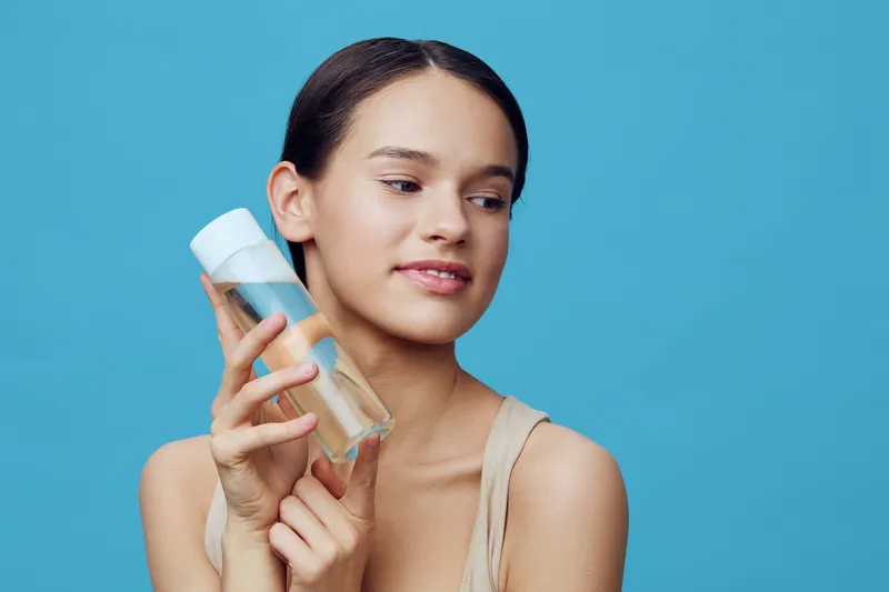 Young woman holding a skincare product, showcasing healthy skin and a radiant smile against a blue background, representing beauty and self care