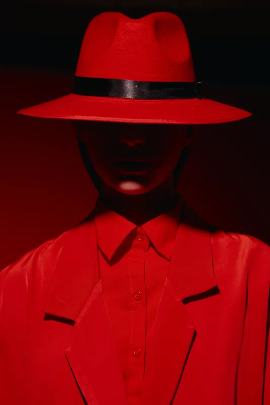 Mysterious female model in a bold red fedora and tailored jacket blends into dark red background. Dramatic shadows hide facial details. Concept for fashion, beauty, creative style.