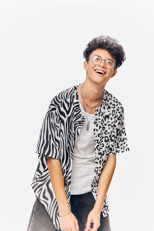 Young man wearing animal print shirt and glasses smiling joyfully, with curly hair and casual style, isolated on white background, modern fashion portrait.