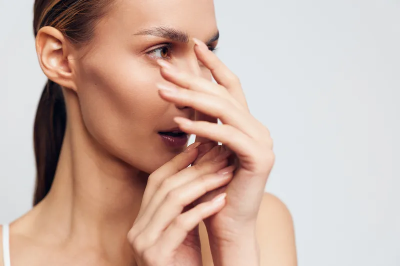 A young woman with clear skin and natural makeup gently touches her face near the nose and mouth, looking to the side against a plain white background. Concept for skincare and beauty.