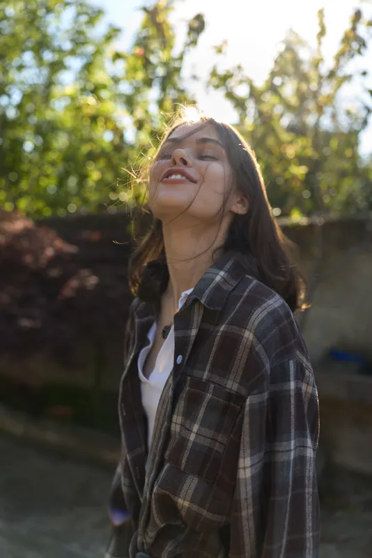 A relaxed woman enjoying outdoor lifestyle, smiling in a plaid shirt bathed in warm sunlight. A casual moment captured in a sunlit setting, conveying happiness and freedom