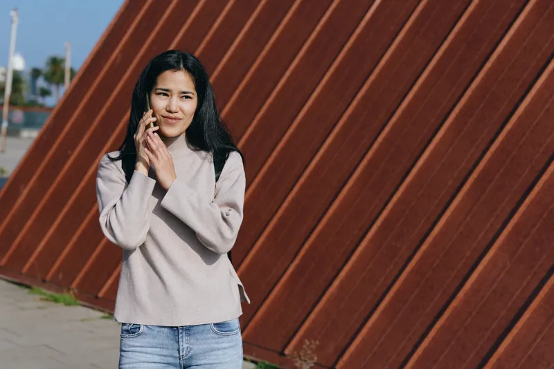 Woman talking on phone outdoors, casual sweater and jeans, smiling near rusted metal wall panels, sunny street scene, candid lifestyle moment, relaxed communication, everyday connectivity