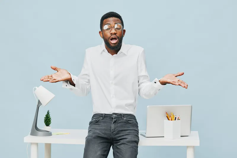 Confused young man with glasses wearing white shirt shrugging shoulders expressing uncertainty in modern office setting isolated on light blue background. People lifestyle concept, casual, emotion, re