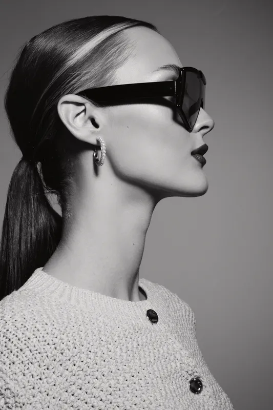 Profile portrait of a pale white woman wearing dark sunglasses and knit sweater. Elegant confident female model with smooth ponytail in studio closeup.