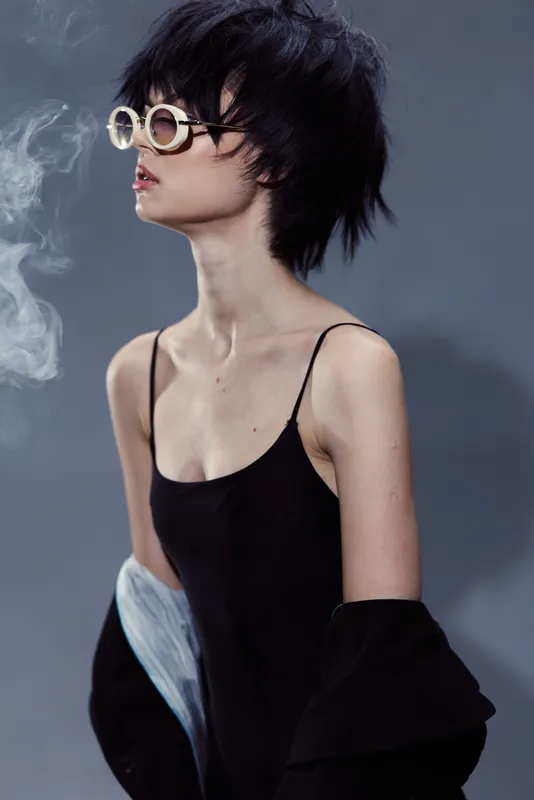 Stylish woman wearing unique round sunglasses and black outfit with smoky background expressing edgy fashion attitude.