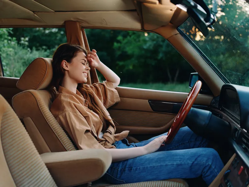 Young woman enjoying a moment of relaxation in a vintage car, wearing a brown oversized shirt and jeans, surrounded by green nature, conveying a sense of freedom and tranquility.