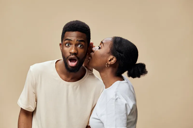 Surprised young African American man listening to a secret from a smiling woman with an astonished expression, in a neutral toned background