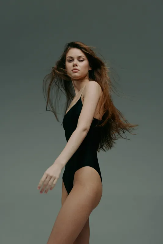 Young woman in black bodysuit posing with flowing hair on gray background, quiet confidence, minimalistic style