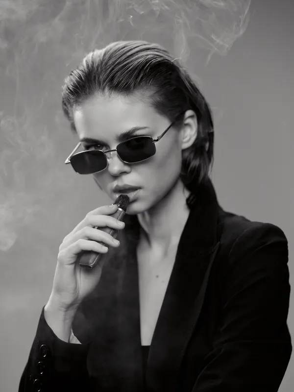Stylish woman in sunglasses vaping with smoke in black and white portrait, wearing dark clothing and slicked-back hair.