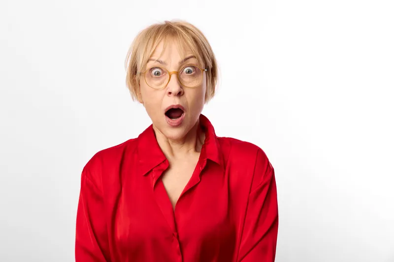 Surprised woman with short blond hair and glasses in a red shirt, shock and disbelief emotion, portrait with open mouth expression on white background