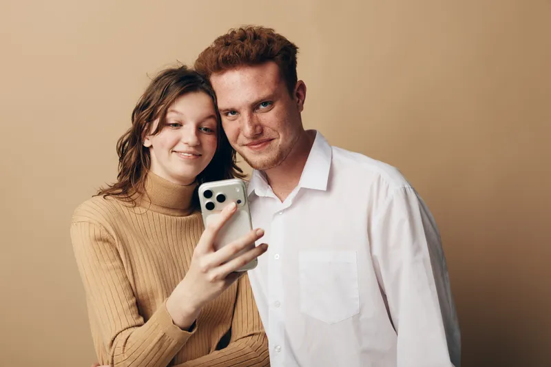 Young couple takes a selfie together using a smartphone. They smile gently, standing close against a plain beige background. Concept of relationships, technology, and casual lifestyle.