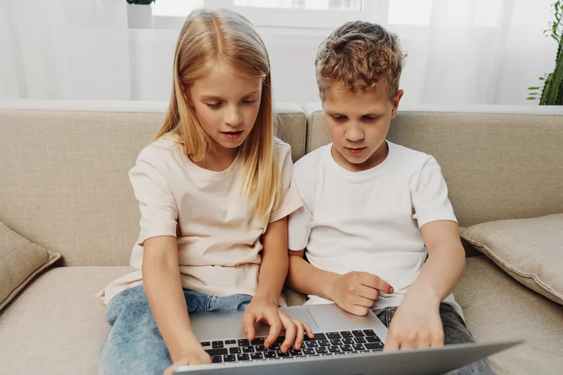 Two focused children sitting on a sofa using laptop together. They wear casual clothes and engage in online learning, digital entertainment, and collaborative activities at home.