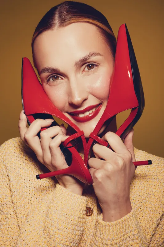 Smiling woman holds red high heel shoes near face, wearing yellow sweater. Studio portrait with warm lighting on brown background. Concept of fashion, style, and happiness.