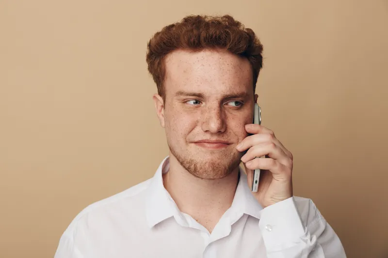 Young man with red hair and freckles wearing white shirt talks on smartphone. He looks sideways with slight smile on beige background. Concept of communication, technology, and casual lifestyle.