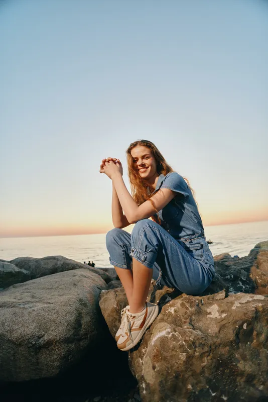 smiling woman sitting on rocks by the sea wearing casual denim jumpsuit and sneakers during golden hour sunset with clear sky and calm ocean background