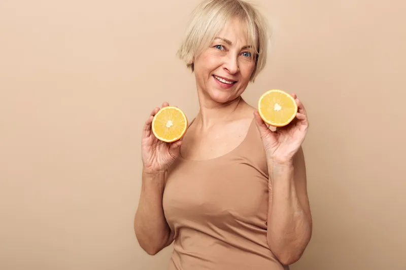 Happy mature woman with short blonde hair holds two halves of fresh orange fruit near face on beige background. Concept of healthy lifestyle, nutrition, and natural vitamins for seniors.