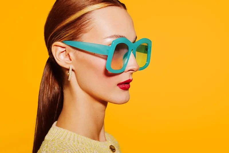 Stylish woman with sleek hair wears oversized turquoise sunglasses and red lipstick against bright yellow background. Fashion portrait with vibrant colors and modern accessories.