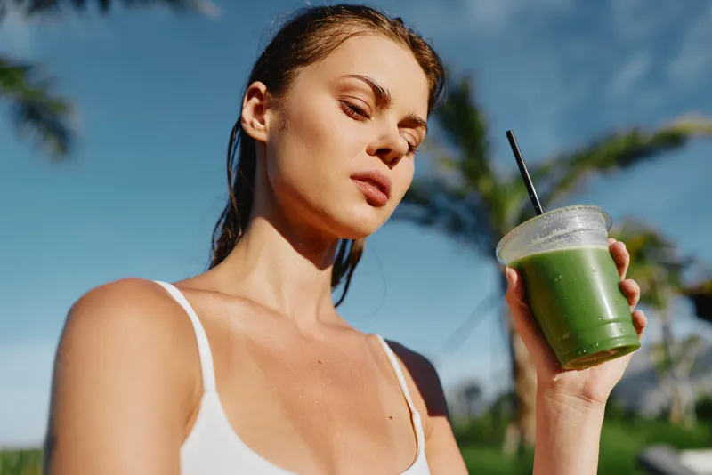 A woman in a white top holds a green smoothie outdoors, basking in bright sunlight. The scene conveys a casual, healthy lifestyle and refreshment during a sunny day.