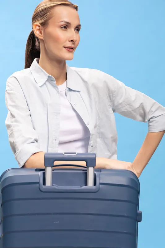Young woman standing with a large suitcase in front of a blue background. Casual travel concept with a thoughtful female traveler dressed in a white shirt and light jacket.