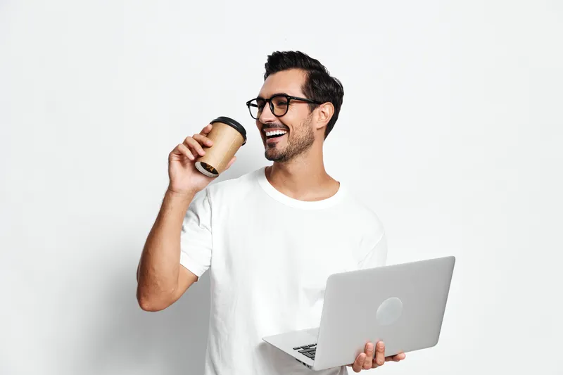 Happy smiling man holding a takeaway coffee cup while working on his laptop, dressed casually in a white t-shirt, isolated on plain solid background. People lifestyle concept.