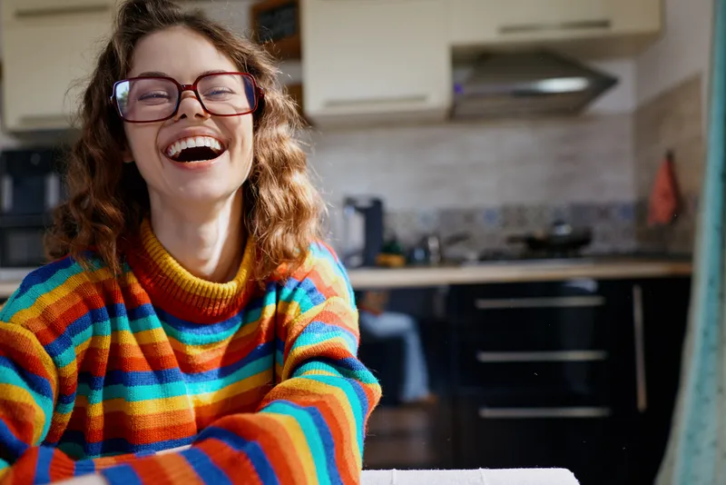 Joyful woman wearing a colorful striped sweater laughs in a bright home kitchen, a candid lifestyle moment, warm light, relaxed mood, everyday happiness, feminine charm, friendly atmosphere