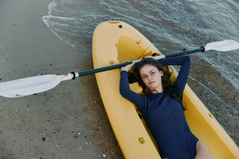 A woman lies inside a bright yellow kayak on a sandy shoreline, resting with a double paddle across her chest while gentle waves lap the hull under soft daylight.