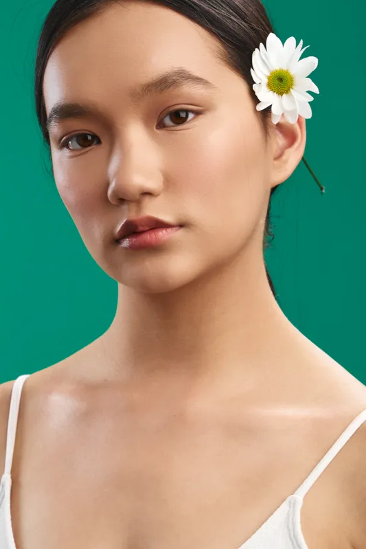 asian woman in a studio portrait with soft lighting and a white flower behind her ear, conveying calm confidence, natural beauty, and quiet focus in a minimalist scene.