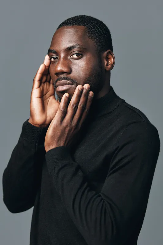 African man with dark skin wearing black sweater posing with hands on face in creative conceptual visual style studio portrait. Serious expression and thoughtful mood on gray background.