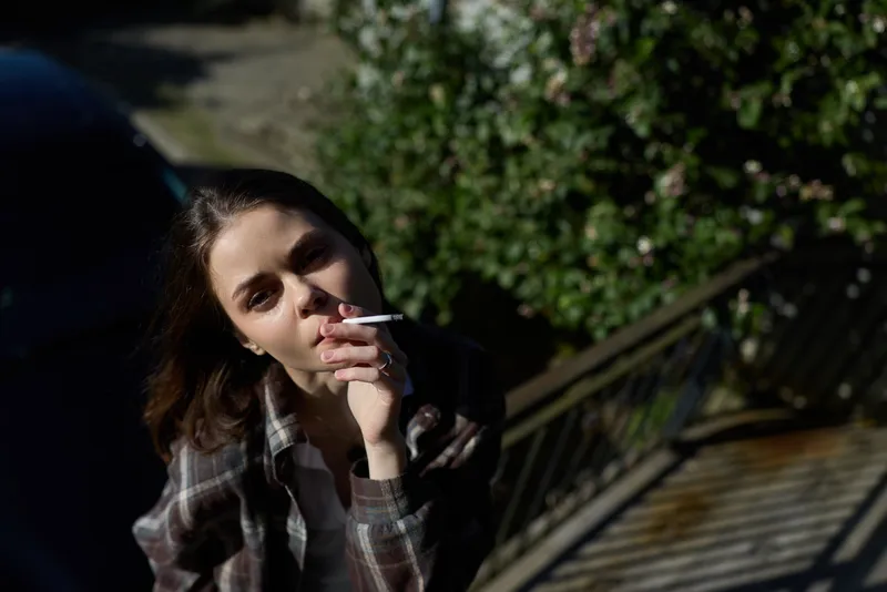 A woman in a plaid shirt sits outdoors, smoking a cigarette, looking pensive. The sunny street and railing provide a calm, introspective mood