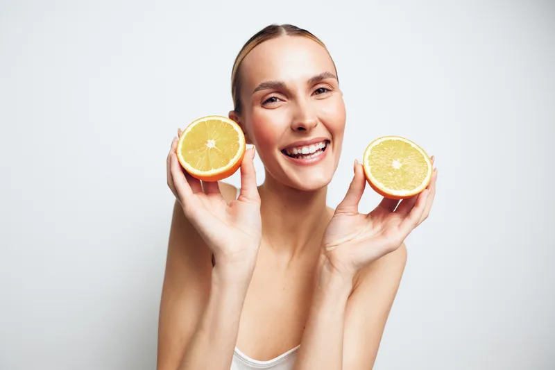 A happy young woman holds two halves of a fresh orange close to her face smiling brightly against a white background. Concept for healthy lifestyle, vitamin C, and natural beauty.