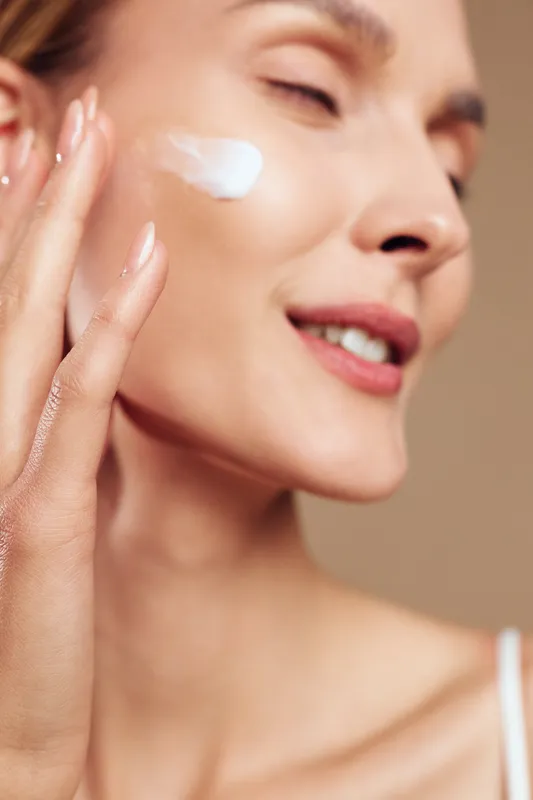 Smiling woman with clear skin applying white face cream on cheek, close-up on beige background. Concept of skincare, beauty routine, wellness, and natural self-care with soft lighting.