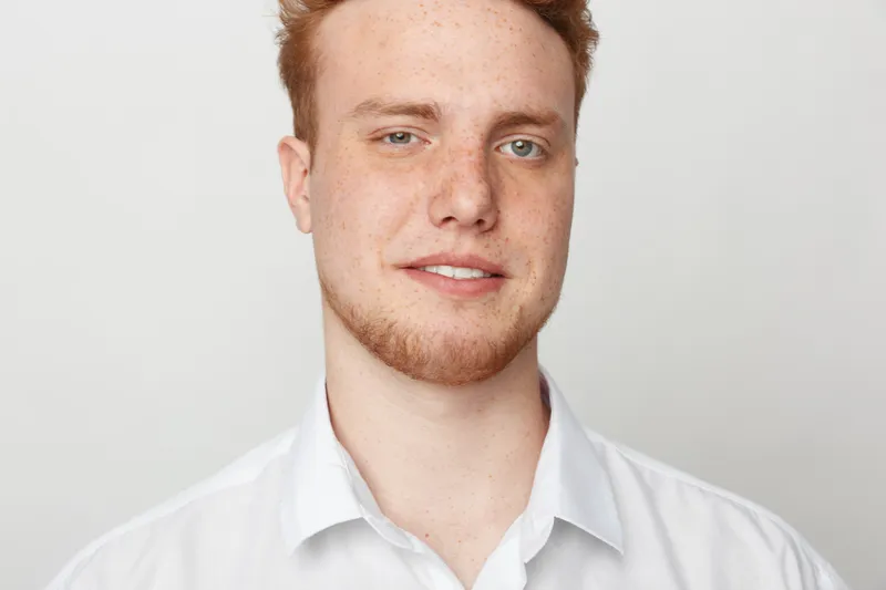 Close-up portrait of a young man with red hair and beard wearing a white shirt smiling gently on a light background. Concept of casual style, confidence, and natural look.