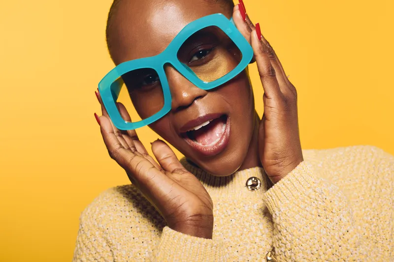 Close-up of a happy dark skinned African woman wearing oversized turquoise glasses and a beige sweater. Bright yellow background creates vibrant, joyful mood for fashion and lifestyle concept.