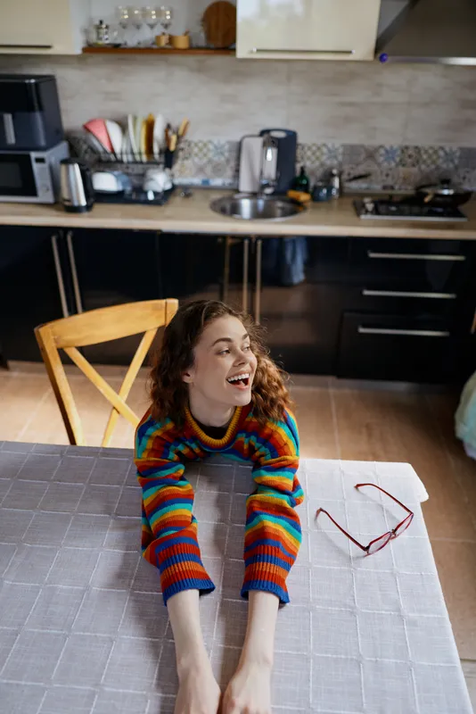 A young woman with curly hair wearing a colorful striped sweater smiles across a bright kitchen table, enjoying a casual home moment as sunlight streams in and sunglasses rest beside a laptop.
