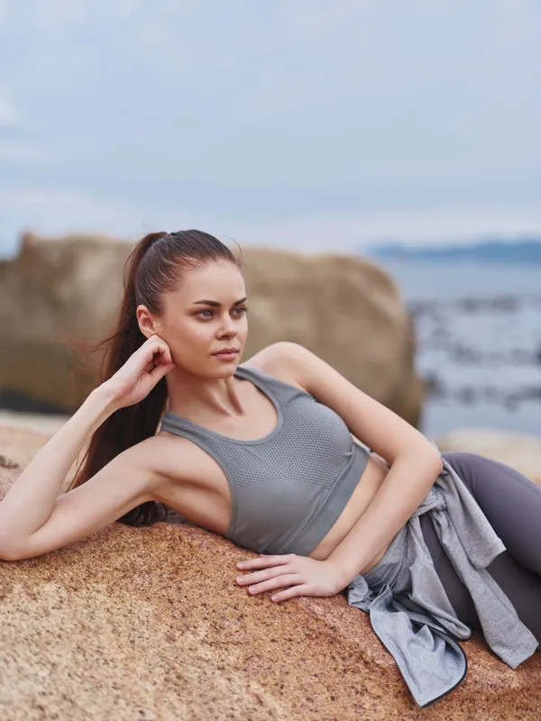 Young athletic woman in gray sportswear lounging by the sea, expressing calmness and strength amidst a natural rocky background during a cloudy day