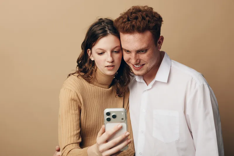 Young couple with smartphone takes selfie together, smiling and looking at screen. Casual clothing and neutral background create warm, intimate moment of connection and technology use.